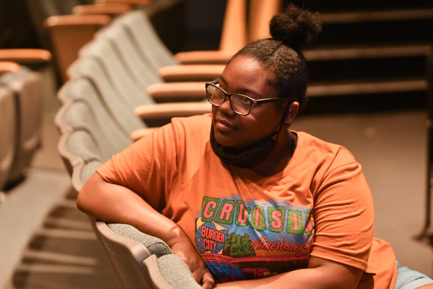 A young woman wearing an orange T-shirt sits in a row of theater chairs listening to someone talking off camera.