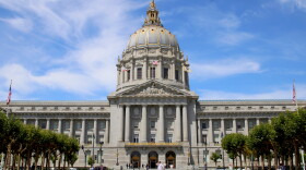 A view of the front of city hall with a few trees in the foreground and a blue sky with wispy clouds behind it. The city hall building is long and rectangular with columns in front, gold trim, and a big dome coming out of the center of it. You can see a few people walking in front of the building. 
