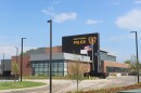 Exterior photo of the new East Peoria Police Department headquarters, a modern two-story building combining red brick, gray metal paneling, and a black accent wall with large illuminated "EAST PEORIA POLICE" lettering and the department's badge-shaped shield logo. Floor-to-ceiling glass windows wrap the lower level, with an American flag, a Thin Blue Line flag, and a city flag flying out front on tall poles.