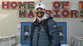A student wearing a face mask, a baseball hat and a backpack stands in front of a school building.