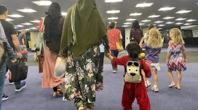 Two Afghan women are seen from behind, wearing muted colored hijabs and long dresses. One woman holds the hand of a small child in a red outfit wearing a dog backpack.