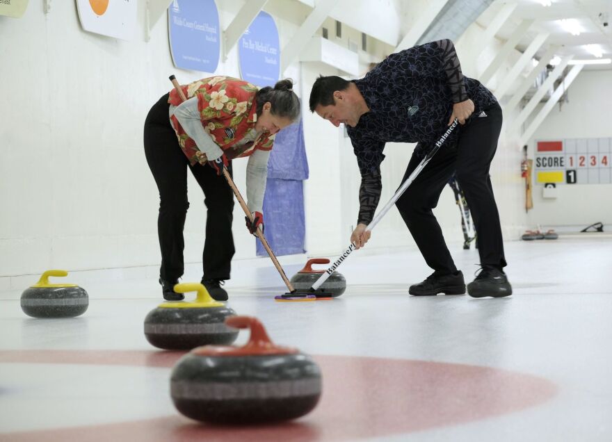 A woman and man curling, holding sticks with stones on the ice