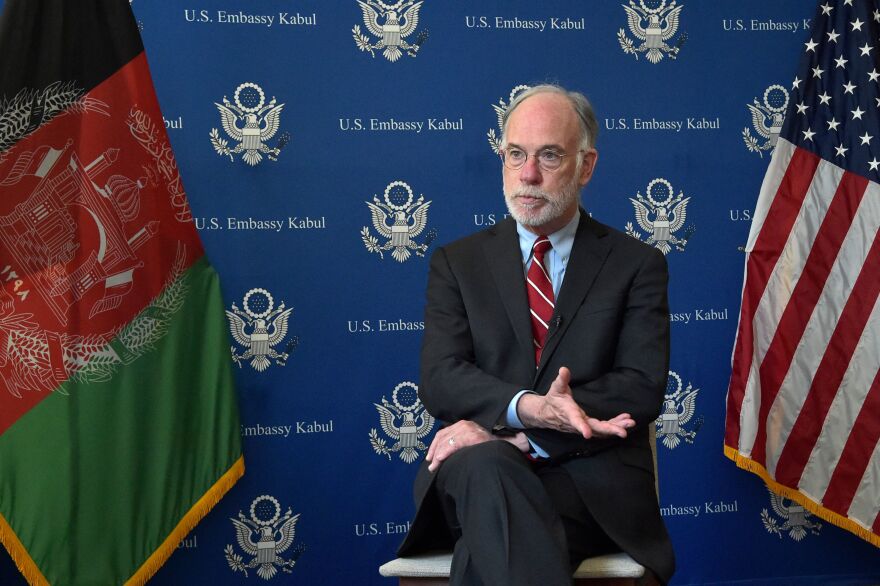 U.S. Ambassador Ross Wilson sits with arms and legs crossed. Behind him are U.S. and Afghan flags and a backdrop reading "U.S. Embassy Kabul".