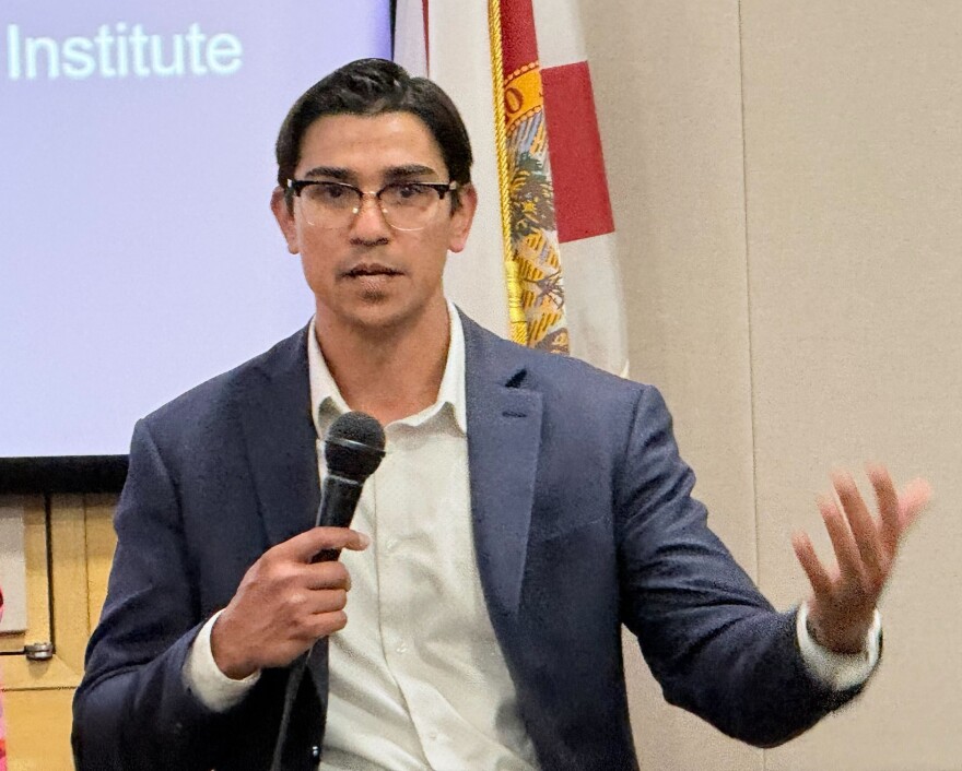 Man with glasses wearing gray sports coat holds a microphone and speaks 