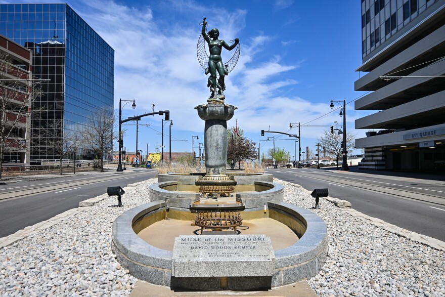 A statue of a nude woman sits atop a pedestal that lies above three circular, granite pools. It is a fountain but it does not have water in it. The figure on the fountain is holding a net and fish mingle around her legs. The fountain is empty.