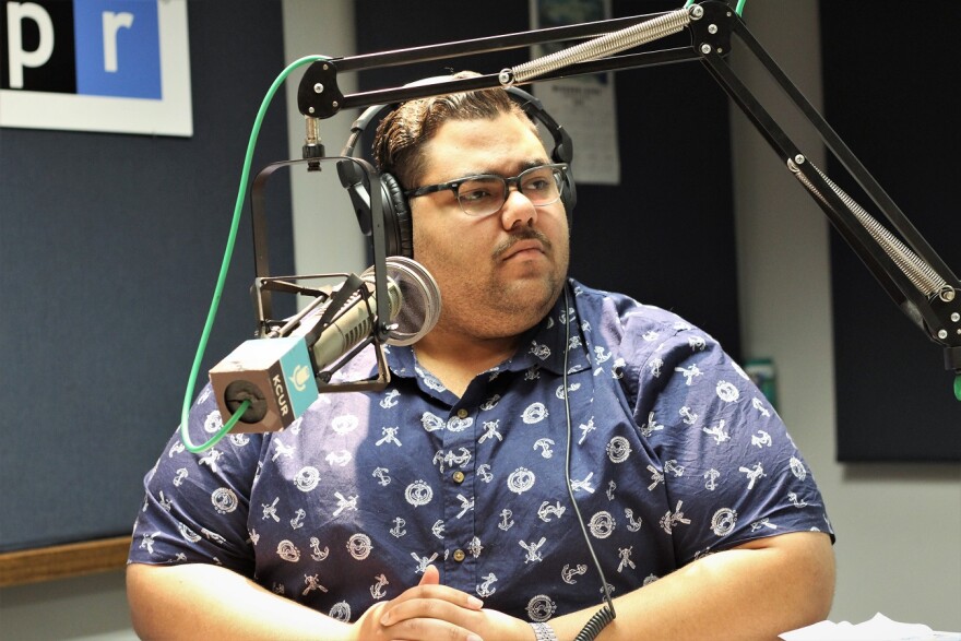 Male in short-sleeved blue shirt wearing headphones and seated in front of a microphone in the KCUR studio.