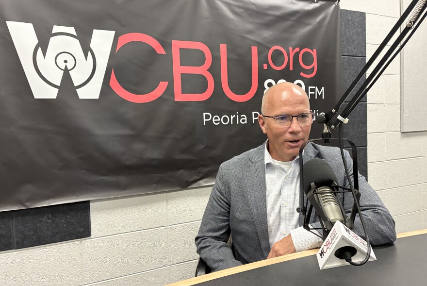 A man in a gray blazer and white shirt speaks into a microphone in a radio studio with a WCBU.org 89.9 FM banner behind him.