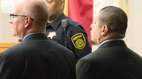 Two men in suits look offscreen. A sheriff's deputy stands behind them.