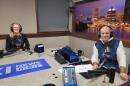 Two people wearing headphones sit at a table in a radio talk studio: a woman at left has long blonde hair and is wearing glasses and a long-sleeved black shirt; a man at right has short dark hair and is wearing a puffy navy vest over a white shirt.