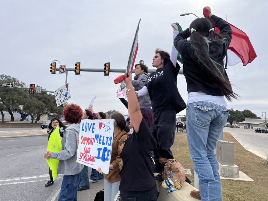 Students stand on the barrier at the edge of an intersection carrying signs that say "Love Melts ICE. Support our Immigrants" and "Skipping Lessons to Teach One."