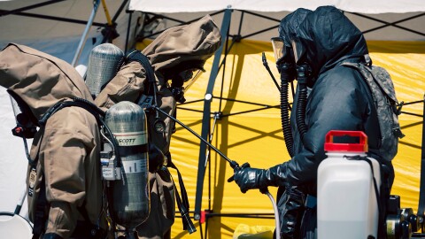 Two people in emergency response suits and masks walk past tents. 