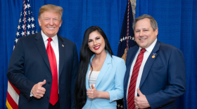 Photo shows three people who have one thumb up each. There is a taller man in a navy blue suit and red tie on the right. In the middle is a pretty brunette with long dark hair wearing a powder blue suit and a man on the right in a red, blue, and coral diagonal-striped tie. He is also wearing a blue suit. All three are smiling and look like they are enjoying themselves.