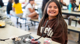 Una niña mira a la cámara con una gran sonrisa, junto a un plato con fruta y otros alimentos.