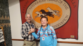 Two men stand in front of the CTUIR flag clasping hands and smiling toward the camera. The man on the left is white, tall, wearing a plaid shirt and khaki pants, the man on the right is shorter, Native, his hair braided into two braids, pulled over his shoulder, he's wearing a blue patterned shirts with geometric, patterns and jeans.