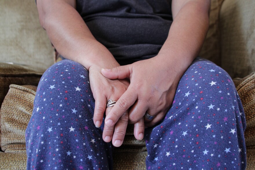 A Nicaraguan woman, above, and her family came to the United States from Costa Rica after experiencing violence in there. They arrived in the Kansas City area in 2024 and are seeking asylum. The woman is sitting on a light brown couch. The photo is cropped so her face is not available. She wears blue pants with red, white and blue stars. Her hands are placed together on her lap.