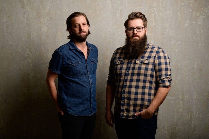  Two men, one with a closely cropped beard and another with a longer beard down to his chest, look at the camera with serious expressions while standing against a concrete background.