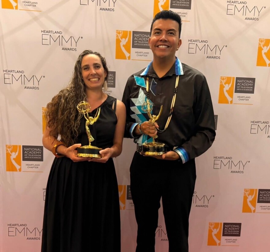 Two dressed-up people hold gold statues and smile at the camera, in front of a backdrop with the words “Heartland Emmy Awards” repeated across the background.