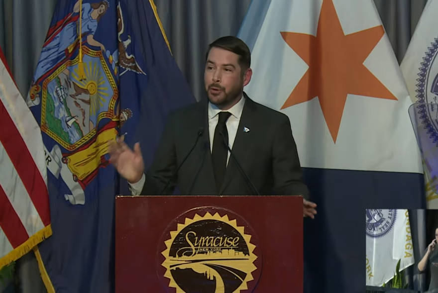 A man in a dark gray suit stands behind a lectern with the American, New York State, and City of Syracuse flags behind him.