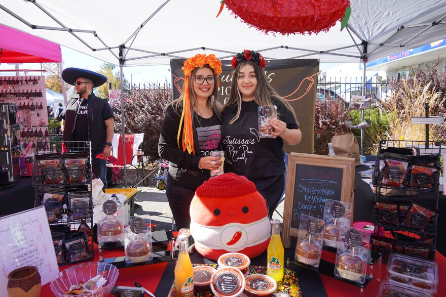 A mother and daughter are smiling while standing behind a booth featuring their products for sale.