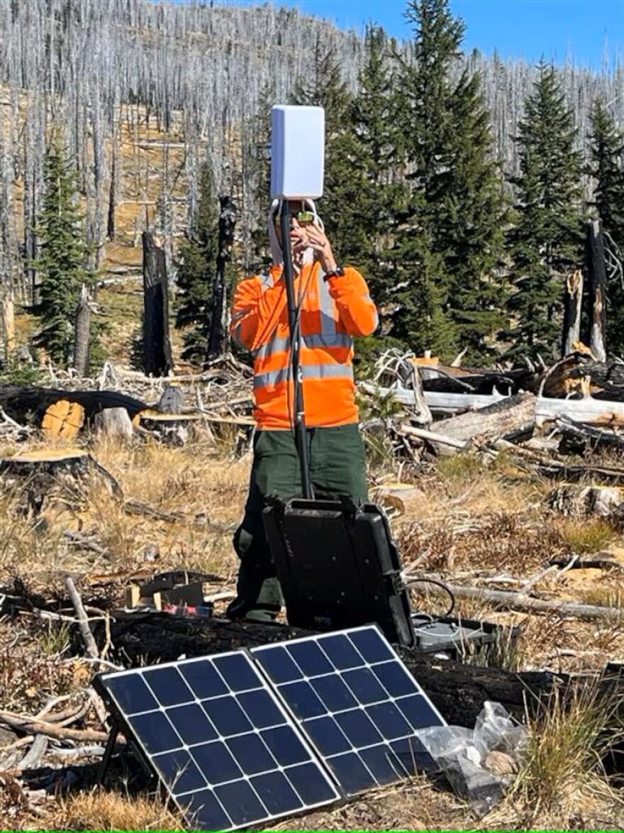 A man in an orange shirt with reflective patches uses an electronic device while looking off into the distance with sunglasses on.