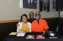 Three people sit smiling at a table covered with copies of a paperback book. A woman in a yellow top and white floral cardigan sits on the left, a man in a black shirt and glasses stands behind, and an older man in a bright orange shirt and sunglasses sits on the right.