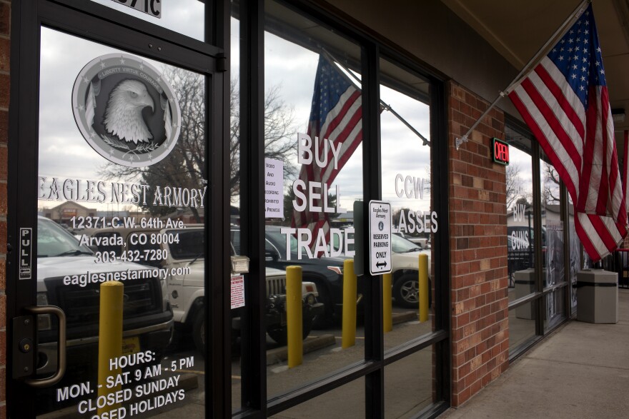 The Eagles Nest Armory gun shop is seen on March 24, 2021, in Arvada, CO. This shows a glass windowed storefront of a strip mall where a flag blows in the wind and the signs on the glass say "BUY SELL TRADE"