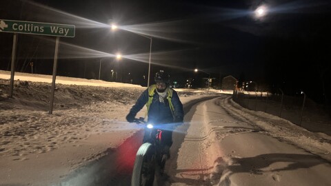 A man in a yellow reflective vest, cold weather clothes, wearing bike helmet rides a fat tire bike down a snowy bike path in the early morning darkness, illuminated by streetlights.