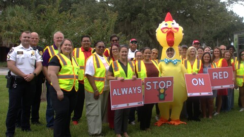 A group of people in neon traffic vests pose for a photo. They hold signs that read, Always Stop On Red. A police officer in uniform and a person wearing an inflatable chicken suit are also part of the group.