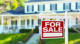 A red FOR SALE sign sits on a lawn in front of a large, white house.