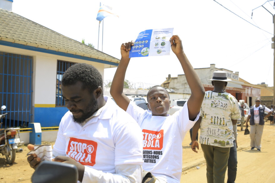 Two African men wearing STOP EBOLA white tshirts walks through the unpaved streets of a village holding up flyers with info about Ebola.  