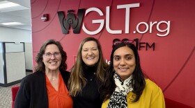 Three women stand shoulder to shoulder smiling at the camera. A large red wall sits behind them in an office. 