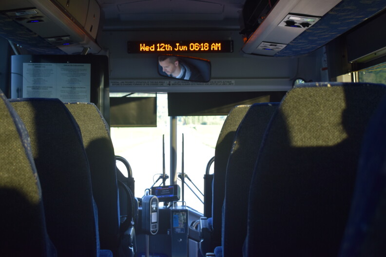 Looking down a bus aisle toward the front where a sign displays "Wed 12th June 6:18 a.m."