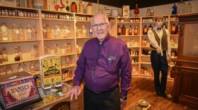 A man wearing a purple shirt rests his hand on a glass cabinet.  Behind him are shelves full of antique pharmaceutical glass containers, a mannequin depicting an old-fashioned pharmacist and other items from an apothecary.