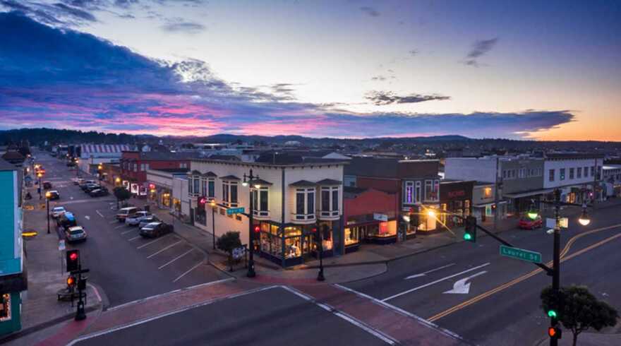 Shops and other buildings line an intersection in downtown Fort Bragg under a colorful evening sky.