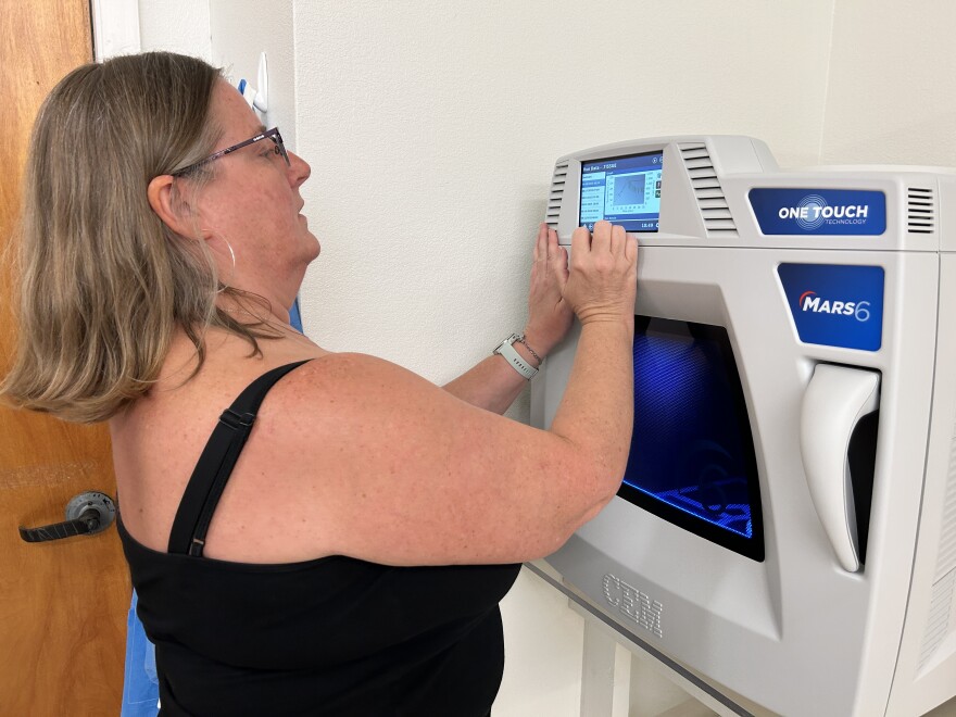 A woman wearing a black tank top and glasses with mid-length dark blond hair looks at a small screen on a machine that resembles a large microwave.