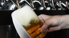Close-up view of a hand holding a glass being filled with honey-colored beer, with foam pouring over the side of the glass