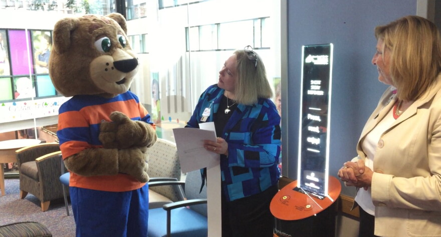 Akron Summit County Public Library mascot "Paws" displays the first story from the new dispenser at Akron Children's Hospital to hospital President Grace Wakulchik (right) and library Director Pam Hickson-Stevenson.