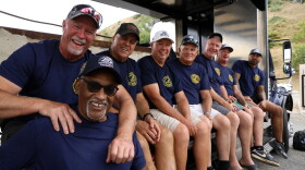 A row of men in blue T-shirts sit on a truck bed