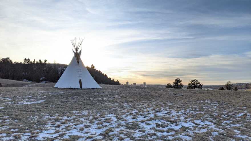 Under a blue sky holding thin clouds that fades to gold at the horizon, stands a white teepee against a backdrop of a cliff and the downtown Billings city outline in the distance. The foreground is a dirt and scattered grass field dusted by light snow.