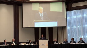 Doug Jones stands behind a podium with people on either side sitting along a table. A screen with his image is above him.