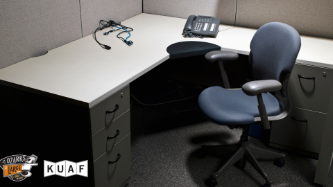 An empty office cubicle with a gray desk, a black office chair, a telephone, and tangled cords.