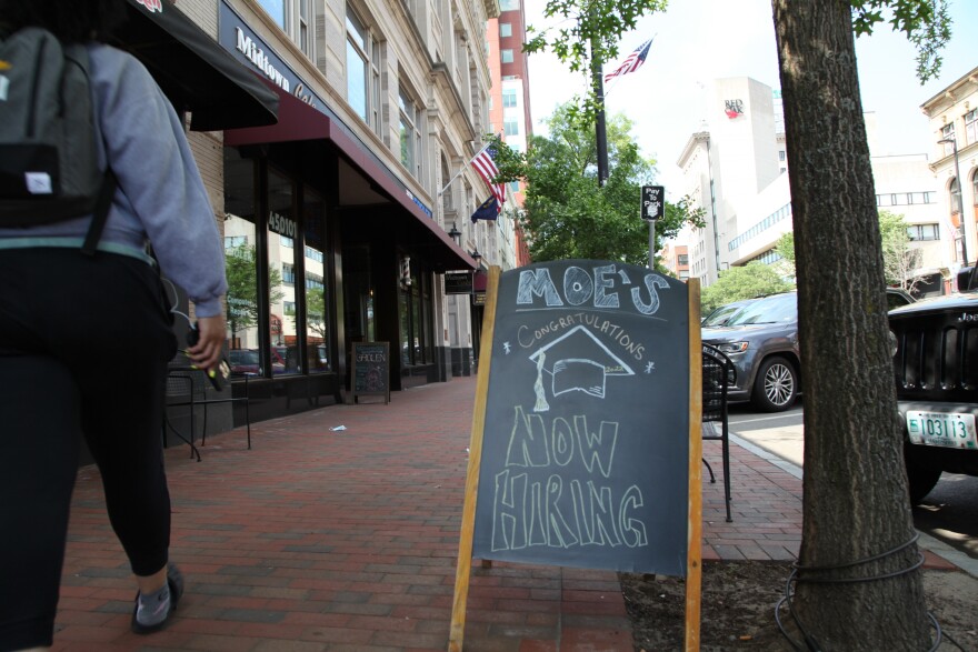 A sign on a sidewalk in Manchester reads: "MOE'S - Congratulations - NOW HIRING" and includes a graduation cap.