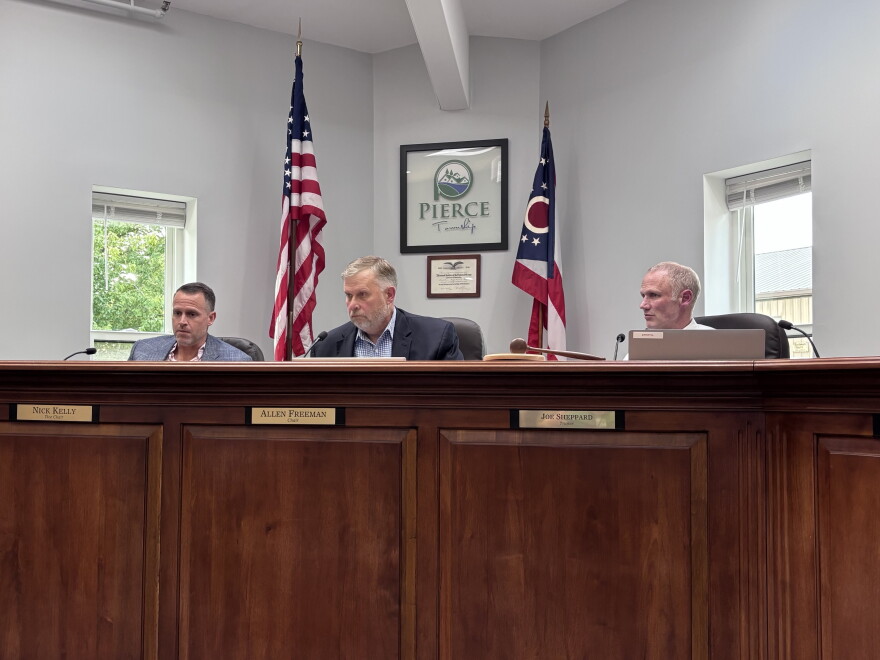 Three men sit behind a large wooden dais, with the Ohio and American flags sitting in the background.