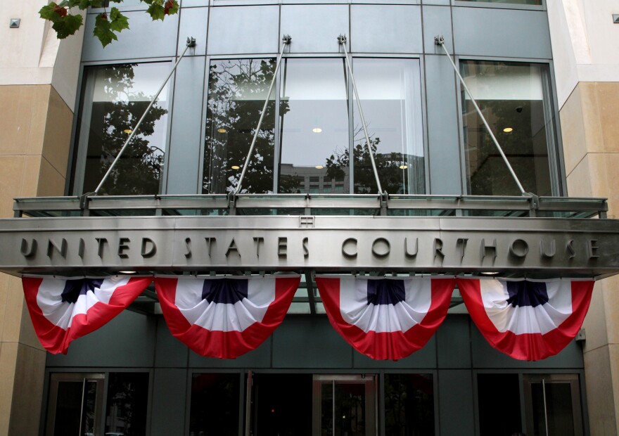 The outside of the Oakland Federal Courthouse is in frame during the day, the words "UNITED STATES COURTHOUSE" are on a plaque in front of the building. There are American flags hanging from the overhang of the building.
