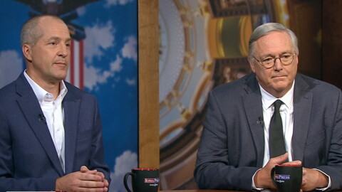 Two men in suits sit a wood desk