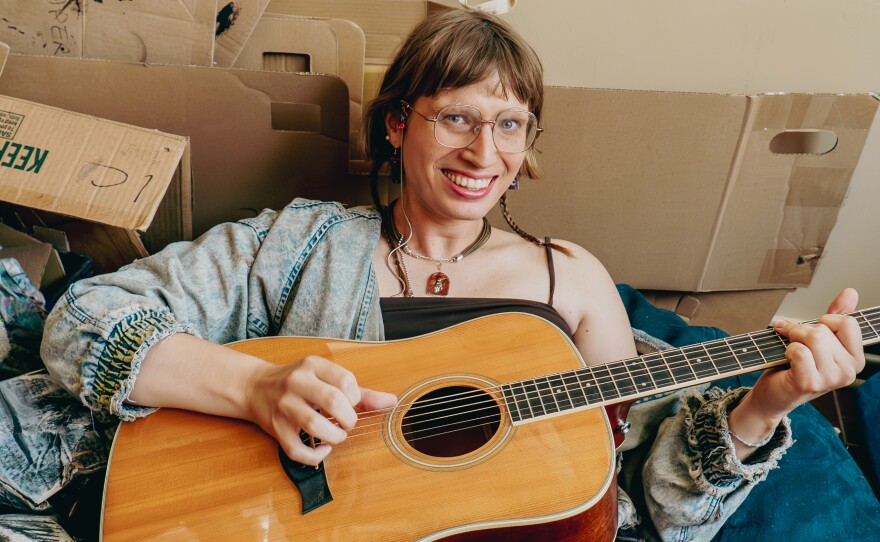 A person with short brown hair wearing glasses, a necklace, and black tank top under an acid wash denim jacket, holding a guitar