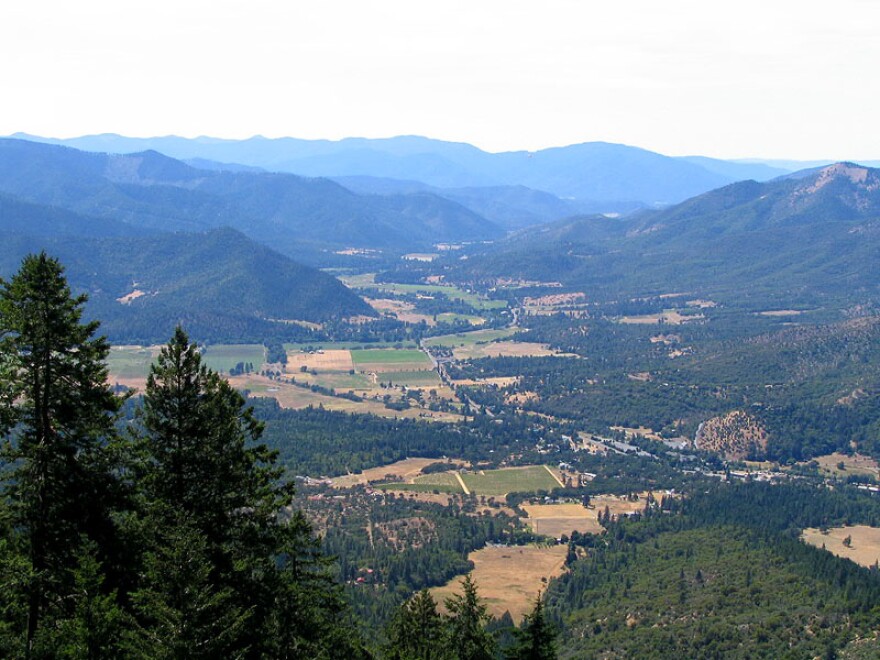 A landscape photo of a valley, surrounded by rolling hills on both sides covered in trees. In the middle is patches of farmland, some roads and structures.