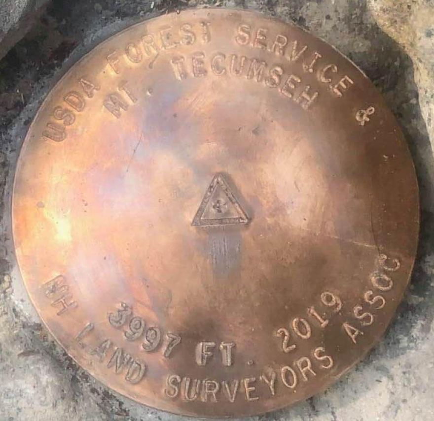 A close-up view of a bronze disc set in granite. It reads: "USDA Forest Service & Mt. Tecumseh. 3997 ft. 2019. NH Land Surveyors Assoc."