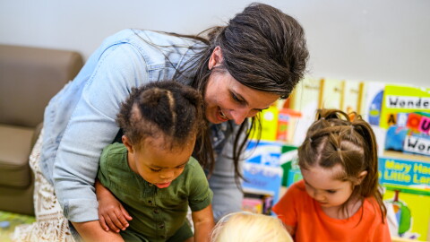 A woman smiles as she interacts with three young children in a classroom, surrounded by colorful books and educational materials.