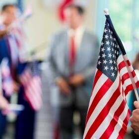 Blurred image of man in suit speaking to a group of people with someone in foreground holding American flag